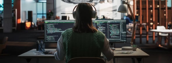 Frau mit Kopfhörern arbeitet an zwei Monitoren mit Code in einem modernen Büro – symbolisch für Cyber Security und Headhunting im Technologiebereich.